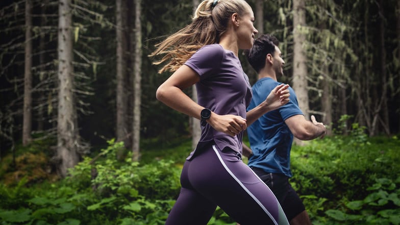 Eine Läuferin und eine Läufer laufen durch den Wald. Beide tragen eine Smartwatch, um ihre Trainingsdaten zu tracken.