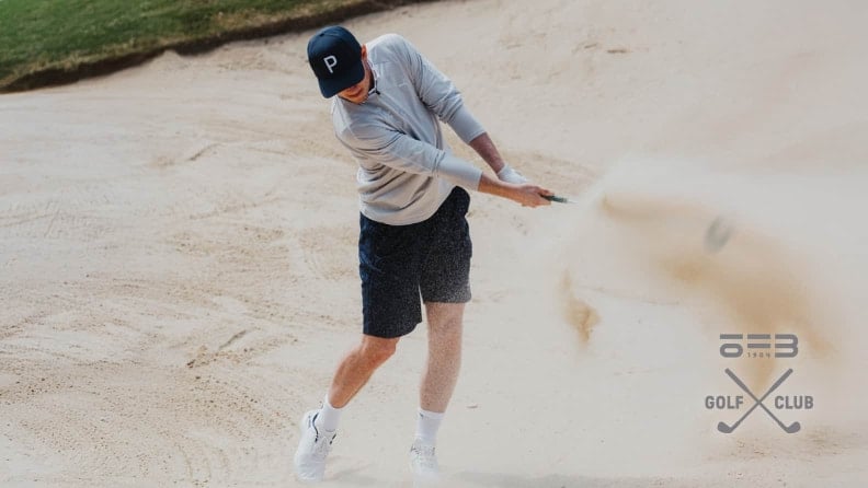 Golfer schlägt einen Ball aus einem Sandbunker und wirbelt dabei Sand auf. Teilansicht des Spielers in Sportkleidung mit Schläger in Bewegung.