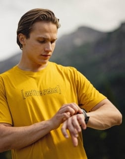 Mann in gelbem T-Shirt blickt auf seine Sportuhr vor einer Bergkulisse.