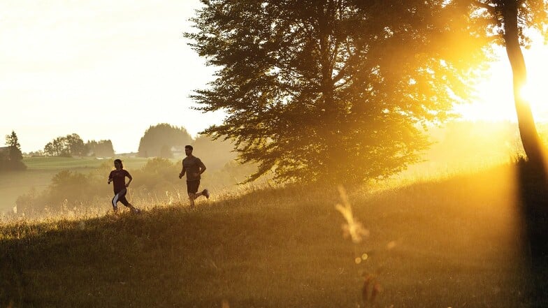 Mann und Frau laufen bei Sonnenaufgang durch eine hügelige Landschaft.