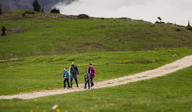 Familie mit zwei Kindern wandert in Wanderkleidung auf einem Schotterweg über eine grüne Almwiese in den österreichischen Bergen