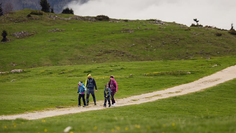 Familie mit zwei Kindern wandert in Wanderkleidung auf einem Schotterweg über eine grüne Almwiese in den österreichischen Bergen