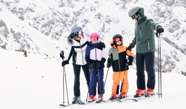 Vierköpfige Familie in Skiausrüstung  auf einer verschneiten Skipiste im Hintergrund eine verschneite Bergkette