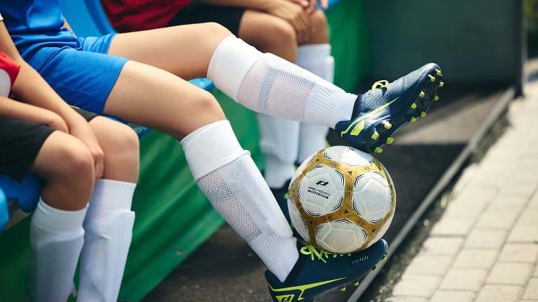 Man sieht die Beine von auf der Ersatzbank sitzenden Kindern. Ein Kind balanciert dabei einen Fußball zwischen den Füßen.