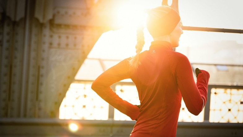 Sportliche Frau joggt bei Sonnenaufgang auf einer Brücke im Gegenlicht.
