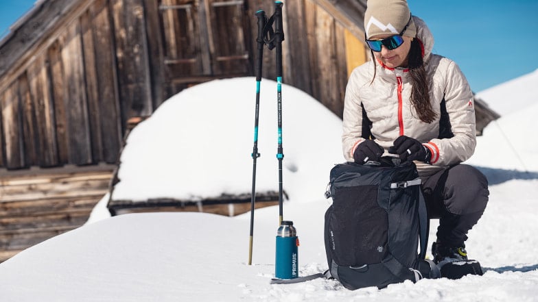 Eine Frau mit Rucksack und Stöcken macht Pause im Schnee.