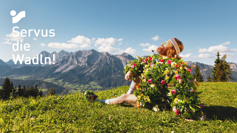 Mann im Blumenoutfit auf Alpenwiese vor Bergpanorama, Servus die Wadln!
