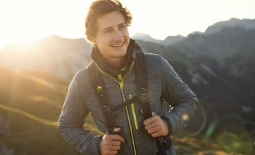 Muž na horách při západu slunce s outdoorovou bundou a batohem.