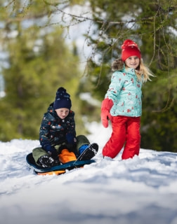 Zwei Kinder spielen im Schnee, eines sitzt auf einem Schlitten, das andere zieht ihn.