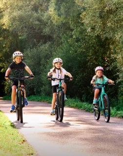 Drei Kinder mit Fahrradhelmen fahren auf einem asphaltierten Weg durch eine grüne Parklandschaft.