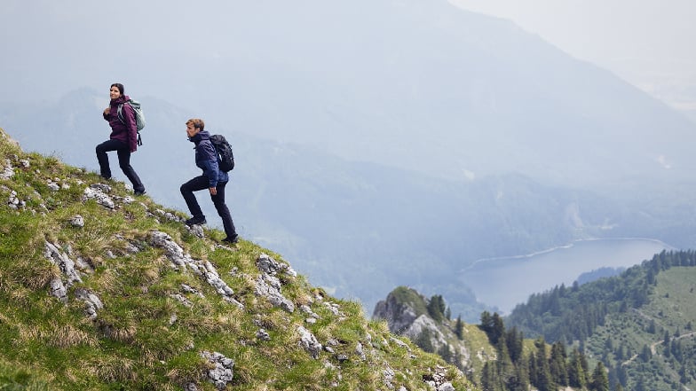 Zwei Wanderer mit Rucksäcken steigen einen steilen, felsigen Grashang hinauf, im Hintergrund ein Bergsee und dunstige Alpenlandschaft