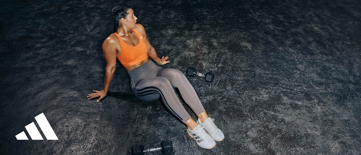 Sportlich gekleidete Frau sitzt auf dem Hallenboden, neben ihr liegen zwei Kurzhanteln.
Im unteren linken Bildbereich ist ein Adidas-Logo zu sehen.