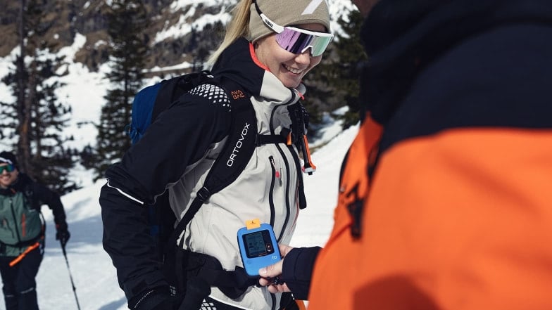 Eine Person zeigt einer lächelnden Frau ein Lawinensuchgerät im Schnee.