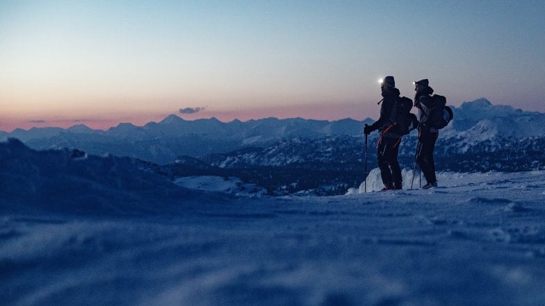 Zwei Skitourengeher im Schnee bei Dämmerung in den Bergen.