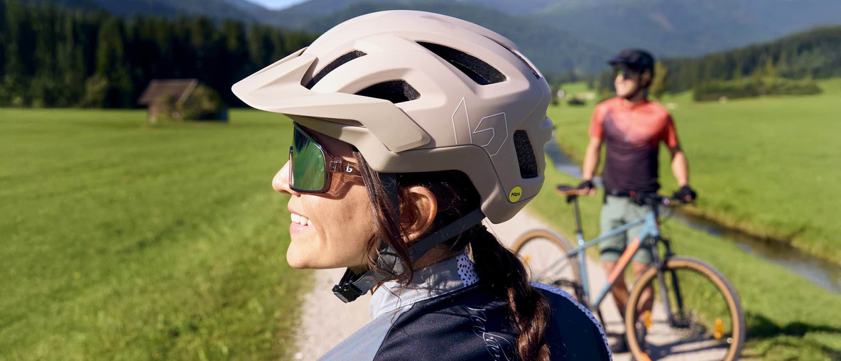 Zwei Personen mit Fahrradhelmen machen auf einem Weg in grüner Berglandschaft Pause.