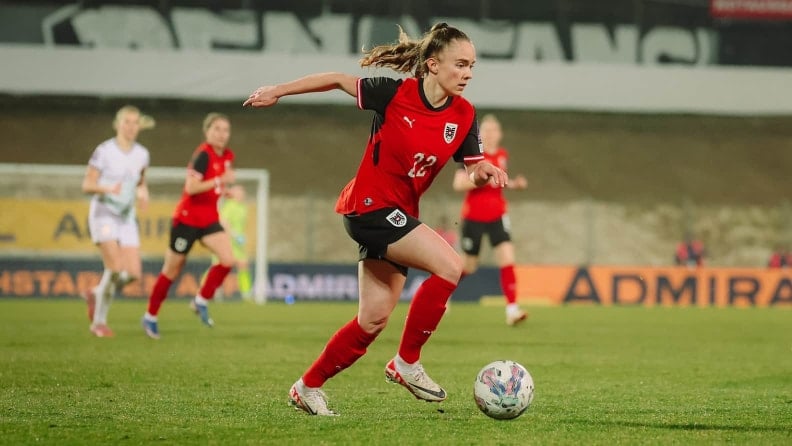 Eine Fußballspielerin in rotem Trikot läuft mit dem Ball über das Spielfeld. Im Hintergrund sind weitere Spielerinnen und ein Tor unscharf zu sehen.