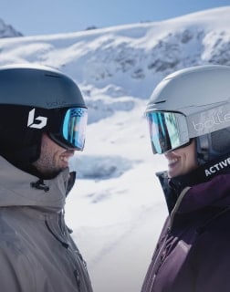 Zwei Personen mit Skihelmen und Skibrillen stehen sich lächelnd gegenüber im Schnee.