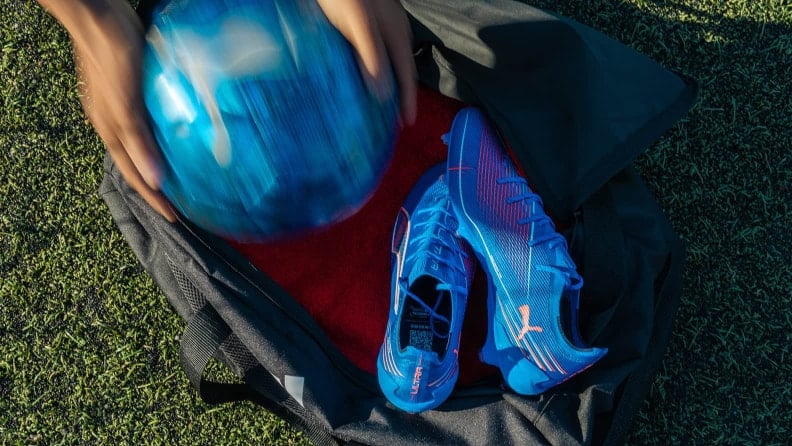Offene Sporttasche auf Rasen mit blauen Fußballschuhen, während ein Ball hineingelegt wird. Sicht von oben auf Ausrüstung und Tasche.