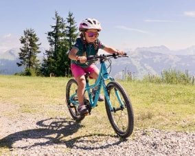 Ein kleines Mädchen fährt konzentriert mit ihrem türkisfarbenen Kinderfahrrad über einen Schotterweg in den Bergen. Sie trägt einen Fahrradhelm und eine Sonnenbrille, im Hintergrund sind Wälder und Berggipfel zu sehen.