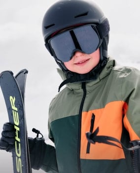 Kind mit Skihelm und Skibrille hält Skier im Schnee.