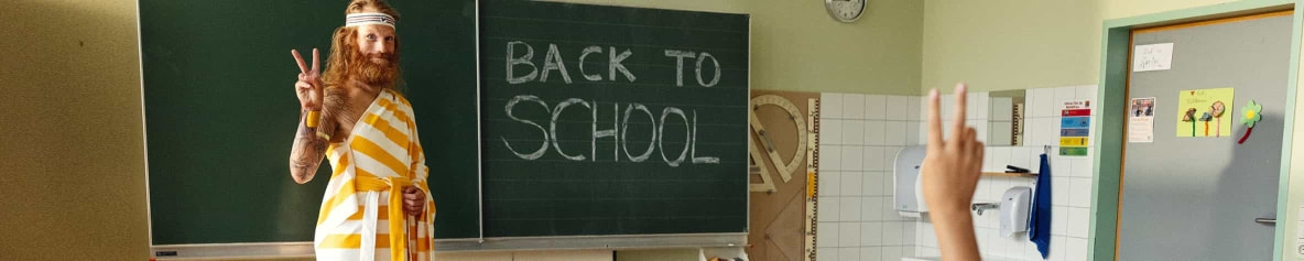Der Sommer steht vor einer dunkelgrünen Schultafel, auf der mit weißer Kreide "Back To School" geschrieben steht und lächelt fröhlich.