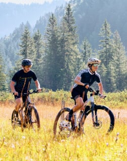 Muž a žena jedou na horském kole po louce s lesem v pozadí.