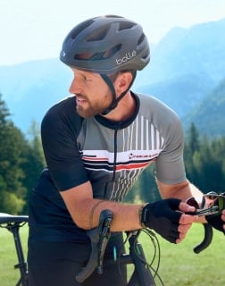Mann mit Fahrradhelm lehnt sich über sein Rennrad und hält eine Sonnenbrille in der Hand, Berglandschaft im Hintergrund.
