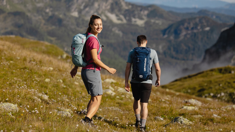 Eine Frau lächelt in die Kamera, vor ihr geht ein Mann mit Rucksack einen Pfad den Berg hinunter.