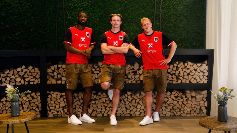 Drei Fußballspieler in roten ÖFB-Trikots und ÖFB-Lederhosen stehen vor gestapeltem Brennholz.