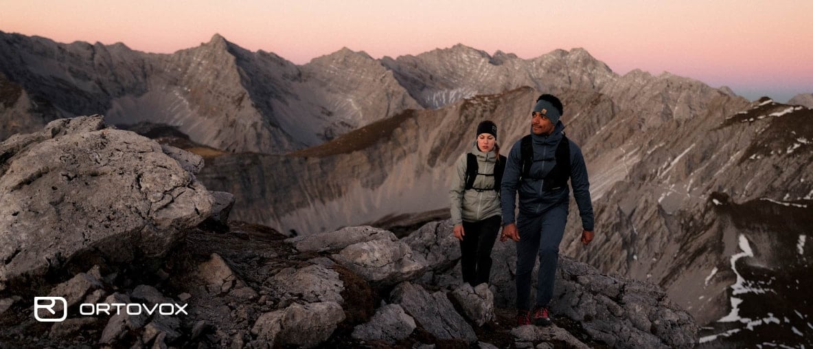 Ortovox-Paar wandert bei Sonnenuntergang über felsiges Hochgebirge mit rosafarbenem Abendhimmel.