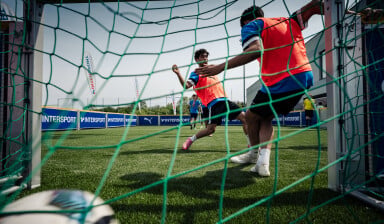 Blick durch das Tornetz auf ein Fußballspiel bei einem INTERSPORT-Event: Zwei Spieler in orangefarbenen Leibchen kämpfen vor dem Tor um den Ball auf Kunstrasen, im Hintergrund sind INTERSPORT-Banden und PUMA-Werbung zu sehen.