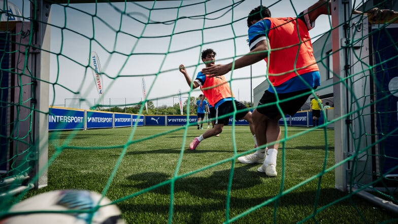 Blick durch das Tornetz auf ein Fußballspiel bei einem INTERSPORT-Event: Zwei Spieler in orangefarbenen Leibchen kämpfen vor dem Tor um den Ball auf Kunstrasen, im Hintergrund sind INTERSPORT-Banden und PUMA-Werbung zu sehen.