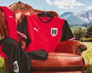 Rot-schwarzes Fußballtrikot hängt auf einem Kleiderbügel an einem braunen Ledersessel im Freien. Daneben liegen passende Kleidungsstücke vor einer Bergkulisse.