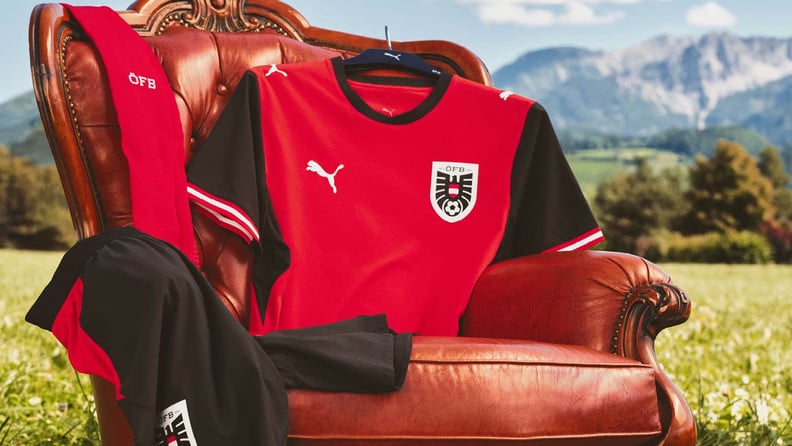 Rot-schwarzes Fußballtrikot hängt auf einem Kleiderbügel an einem braunen Ledersessel im Freien. Daneben liegen passende Kleidungsstücke vor einer Bergkulisse.