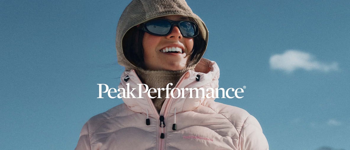 Eine Frau mit einer Jacke von Peak Performance schaut in die Ferne und lächelt. Darüber das Logo von Peak Performance.