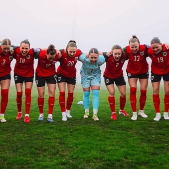 Mehrere Fußballspielerinnen stehen eng nebeneinander mit eingehakten Armen auf dem Spielfeld. Sie beugen sich nach vorne und rufen gemeinsam.