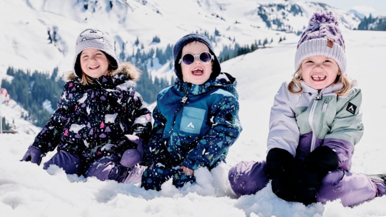 Drei lachende Kinder in Winterkleidung sitzen im Schnee vor einer verschneiten Berglandschaft