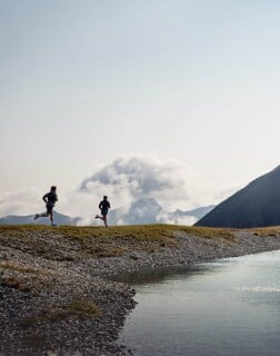 Zwei Personen joggen auf einem Pfad entlang eines Bergsees mit wolkenverhangenen Gipfeln im Hintergrund.