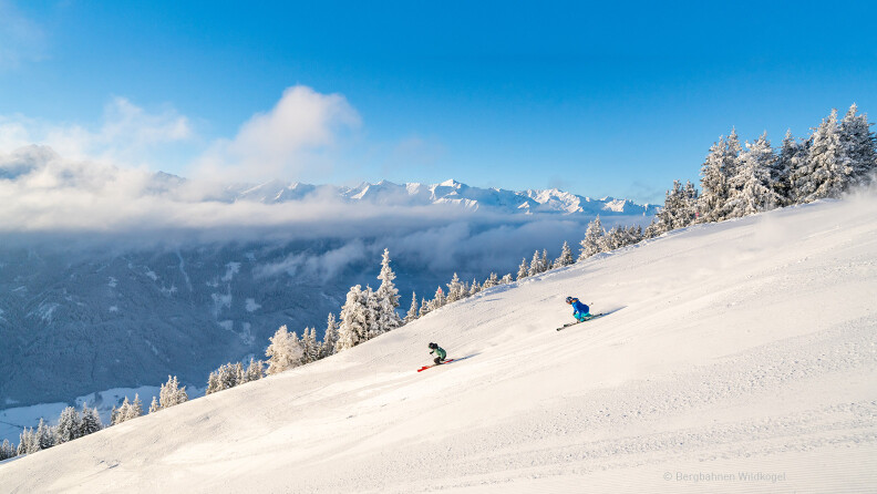Zwei Skifahrer auf sonniger Piste vor einer verschneiten Bergkulisse.