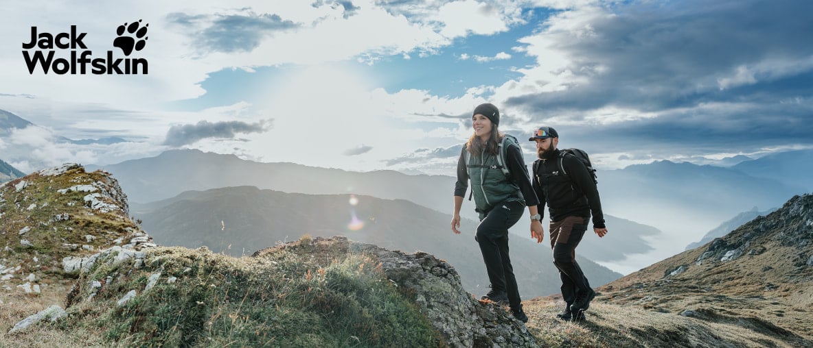 Zwei Wanderer auf einem Berggrat mit weitem Alpenpanorama unter dramatischem Himmel