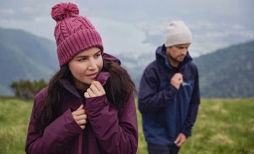 Zwei Personen in warmer Outdoor-Kleidung wandern auf einer grünen Bergwiese mit Blick auf ein Tal und Wasser im Hintergrund.