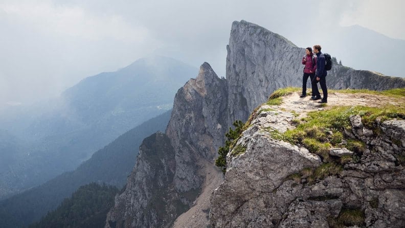 Zwei Wanderer stehen mit Rucksäcken auf einem Felsplateau mit Blick auf ein beeindruckendes Bergpanorama.