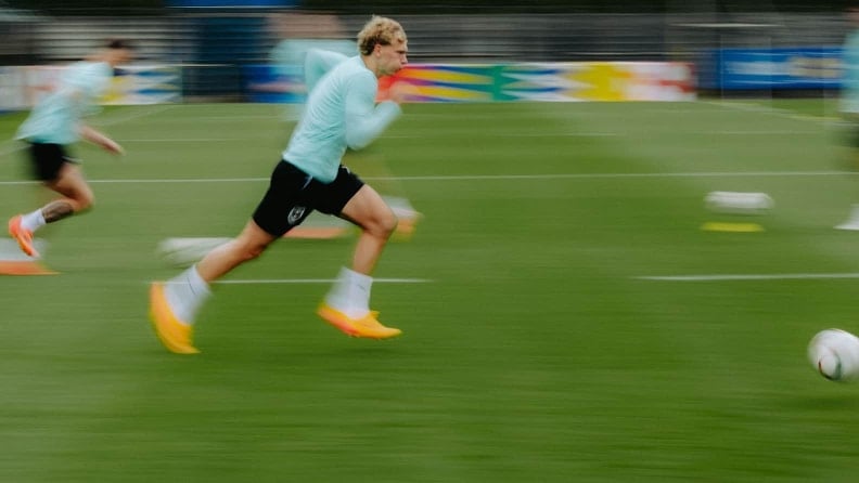 Ein Fußballspieler sprintet im Training über den Rasen, im Hintergrund sind weitere Spieler und Trainingsmaterial unscharf zu sehen.