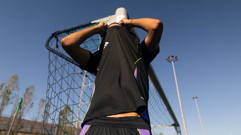 Fußballspieler steht vor einem Tor und zieht sich das Trikot über den Kopf. Das Tornetz und der blaue Himmel sind sichtbar.