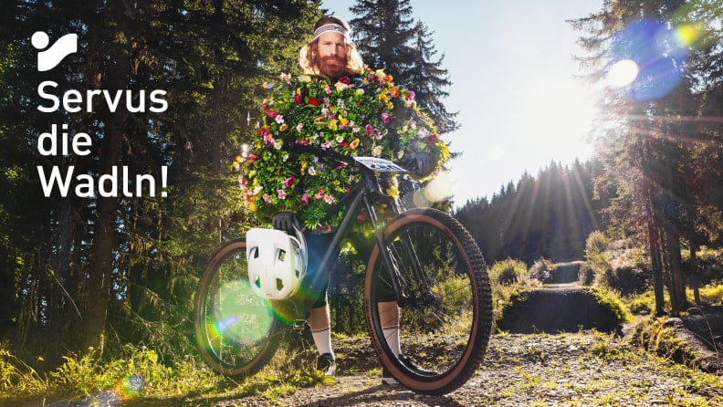 Radfahrer mit Blumenkostüm auf Mountainbike im Wald bei Sonnenlicht, Text „Servus die Wadln!“