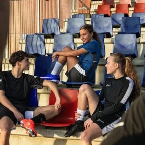 Zwei Fußballerinnen und ein Fußballer erholen sich in der Trainingspause bei einem Gespräch auf der Zuschauertribüne.