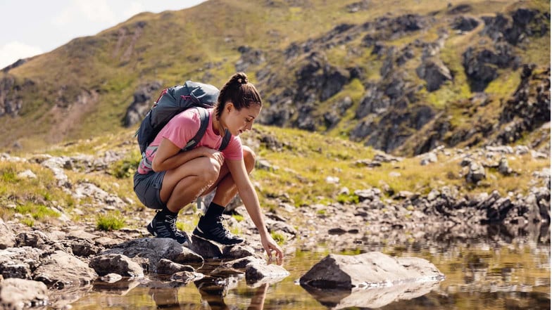 Eine Frau testet die Temperatur eines kleinen Bergsees mit ihren Fingern.