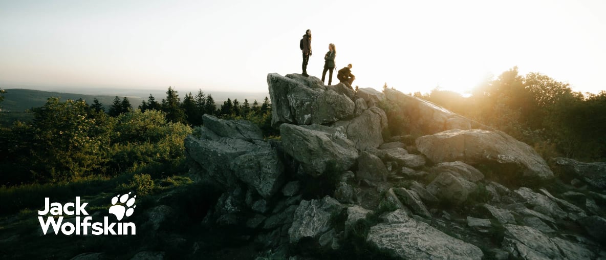 Drei Personen auf einem Felsen in bewaldeter Landschaft bei tief stehender Sonne, Jack-Wolfskin-Logo im Vordergrund