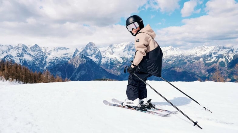 Dítě v lyžařské helmě a brýlích na svahu s výhledem na zasněžené hory, připravené na jízdu.