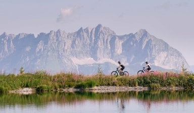 Zwei Mountainbiker mit E-Mountainbikes an einem See vor einer beeindruckenden Bergkulisse.